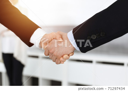 Businessman and woman shaking hands with colleagues at the background. Handshake at meeting in sunny office Businessman and woman shaking hands with colleagues at the background. Handshake at meeting in sunny office 71297434