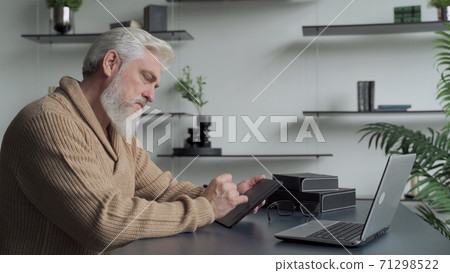 An elderly man with a gray beard draws with a pen on a tablet at home in the living room 71298522