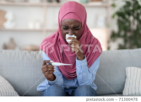 Sick black muslim woman with runny nose checking body temperature at home 71300478