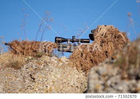 sniper in ghillie (camouflage) suit with a high-precision rifle with optic scope in a combat position in the mountains 71302870