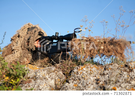 sniper in ghillie (camouflage) suit with a high-precision rifle with optic scope in a combat position in the mountains 71302871
