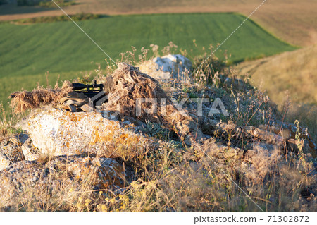 sniper in ghillie (camouflage) suit with a high-precision rifle with optic scope in a combat position in the mountains 71302872