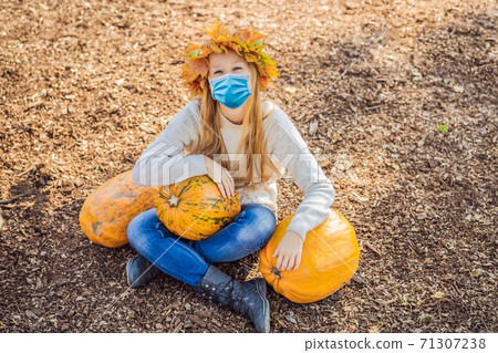 Outdoors lifestyle photo of happy woman wearing a medical mask during COVID-19 coronavirus sitting on the farm. Holding a pumpkin. Wearing knitted pullover and wreath of maple leaves. Autumn colors Outdoors lifestyle photo of happy woman wearing a medical mask during COVID-19 coronavirus sitting on the farm. Holding a pumpkin. Wearing knitted pullover and wreath of maple leaves. Autumn colors 71307238