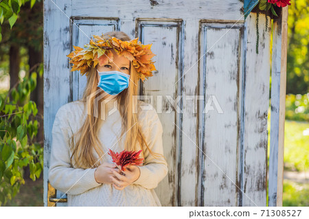 Outdoors lifestyle close up portrait of charming blonde young woman wearing a medical mask during COVID-19 coronavirus wearing a wreath of autumn leaves. Wearing stylish knitted pullover. Wreath of 71308527