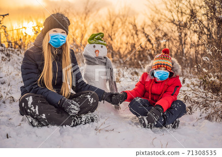 Happy family in warm clothing wearing a medical mask during COVID-19 coronavirus. Smiling mother and son making a snowman outdoor. The concept of winter activities 71308535