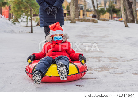 mom son wearing a medical mask during COVID-19 coronavirus ride on an inflatable winter sled tubing. Winter fun for the whole family mom son wearing a medical mask during COVID-19 coronavirus ride on an inflatable winter sled tubing. Winter fun for the whole family 71314644