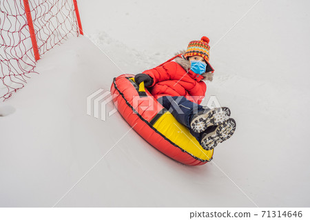 Child wearing a medical mask during COVID-19 coronavirus having fun on snow tube. Boy is riding a tubing. Winter fun for children Child wearing a medical mask during COVID-19 coronavirus having fun on snow tube. Boy is riding a tubing. Winter fun for children 71314646