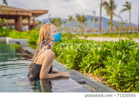 Woman wearing a medical mask during COVID-19 coronavirus relaxing in infinity swimming pool looking at view Woman wearing a medical mask during COVID-19 coronavirus relaxing in infinity swimming pool looking at view 71320593