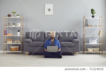Young smiling man sitting on floor with laptop on knees and working online from home Young smiling man sitting on floor with laptop on knees and working online from home 71324638