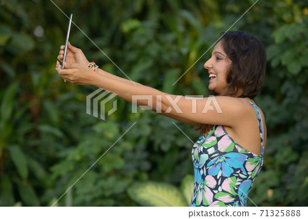Happy young beautiful Indian woman video calling with digital tablet at the park 71325888