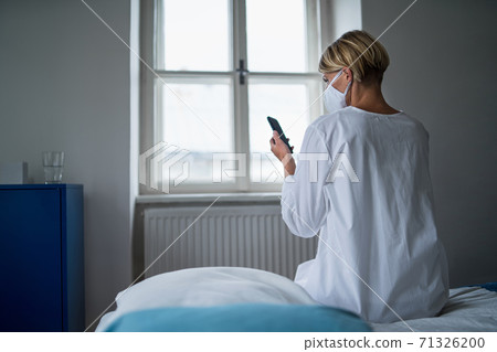 Rear view of woman patient with smartphone sitting on bed in hospital, mental health and coronavirus concept. Rear view of woman patient with smartphone sitting on bed in hospital, mental health and coronavirus concept. 71326200