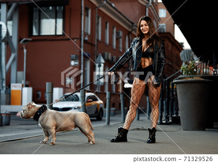 Sexy and srong brunette woman posing with bully dog on city street, looking at camera. 71329526