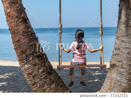 Back view of Asian child girl sitting alone on wooden swing at palm grove, wearing t-shirt and floral legging, enjoying sea view. Back view of Asian child girl sitting alone on wooden swing at palm grove, wearing t-shirt and floral legging, enjoying sea view. 71332721