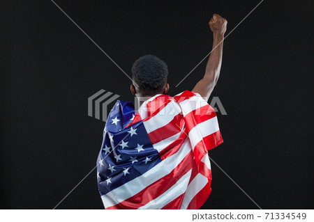 Back view of man wearing american flag standing with raised hand isolated over black background. Concept of patriotism, love for the Motherland, national symbol 71334549