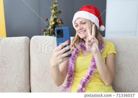 Smiling woman in santa claus hat looks at smartphone and holds her fingers gravely. Smiling woman in santa claus hat looks at smartphone and holds her fingers gravely. 71336037