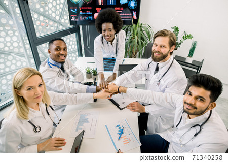 Medicine, healthcare, teamwork and people concept. Multiethnic group of happy smiling diverse doctors holding hands together, while sitting at the table on the meeting at hospital. Top angle view 71340258