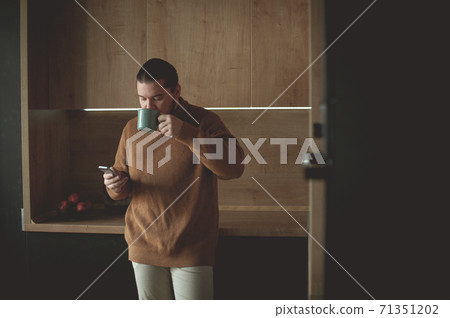 Young man drinks coffee and looks into a smartphone in the kitchen Young man drinks coffee and looks into a smartphone in the kitchen 71351202