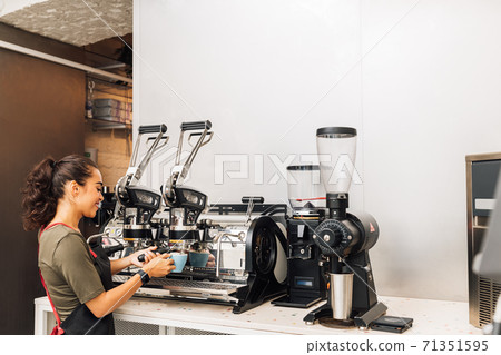 Female barista standing by coffee maker while working in cafe. Woman in apron preparing coffee. Female barista standing by coffee maker while working in cafe. Woman in apron preparing coffee. 71351595