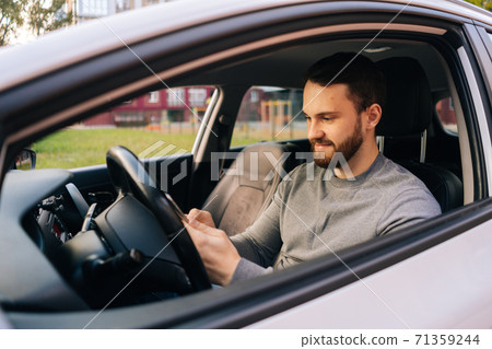 Caucasian bearded young man sitting in car and typing online message on cell phone, side view. 71359244
