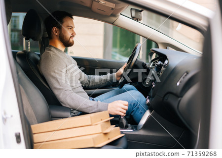 Side view of bearded delivery man wearing casual clothes driving car delivered hot pizza to customer. 71359268