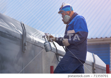 Industrial work. Priming of metal products from the compressor gun. A worker in overalls and a respirator paints the body of a truck trailer or a metal car. Industrial work. Priming of metal products from the compressor gun. A worker in overalls and a respirator paints the body of a truck trailer or a metal car. 71359701