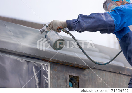 Industrial work. Priming of metal products from the compressor gun. A worker in overalls and a protective mask paints the body of a truck trailer or a metal car. 71359702