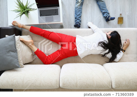 Woman lies on couch next to an alcohol bottle and man lies. 71360389