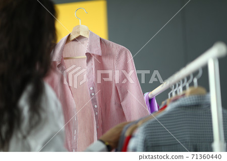 Woman stands with her back next to clothes hanger and holds shirt on hanger in her hands Woman stands with her back next to clothes hanger and holds shirt on hanger in her hands 71360440