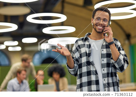 Young happy mixed race man talking by phone and smiling while standing in coworking space with colleagues working on the background Young happy mixed race man talking by phone and smiling while standing in coworking space with colleagues working on the background 71363112