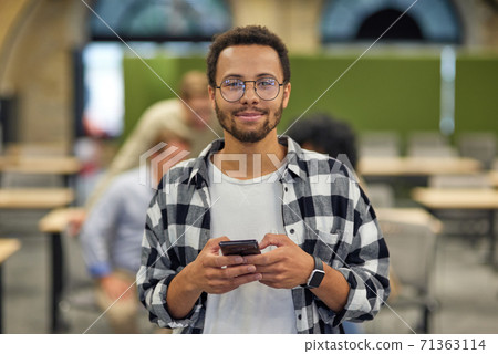 Portrait of a young happy mixed race man holding smartphone and looking at camera while working in the modern office, selective focus Portrait of a young happy mixed race man holding smartphone and looking at camera while working in the modern office, selective focus 71363114