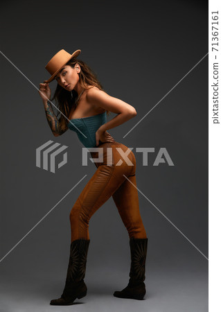 Fashion portrait of young brunette girl in blue western top and jeans with cowboy hat and boots in front of gray copy space studio background. Fashion portrait and lifestyle. Fashion portrait of young brunette girl in blue western top and jeans with cowboy hat and boots in front of gray copy space studio background. Fashion portrait and lifestyle. 71367161