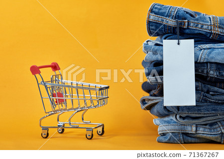 A stack of blue jeans with a white blank tag on a yellow background. Shopping trolley with multi-colored cubes. Sales word written on cubes. Sales consept A stack of blue jeans with a white blank tag on a yellow background. Shopping trolley with multi-colored cubes. Sales word written on cubes. Sales consept 71367267