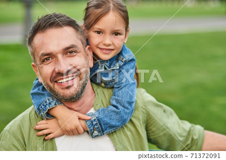 Portrait of young joyful father and his cute little daughter looking at camera and hugging while spending time together outdoors, sitting on a green grass Portrait of young joyful father and his cute little daughter looking at camera and hugging while spending time together outdoors, sitting on a green grass 71372091