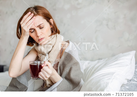 A sick young woman, sitting in bed with a scarf, suffers from a sore throat. Drinking hot tea. A woman with a fever wrapped in a blanket. A sick young woman, sitting in bed with a scarf, suffers from a sore throat. Drinking hot tea. A woman with a fever wrapped in a blanket. 71372105