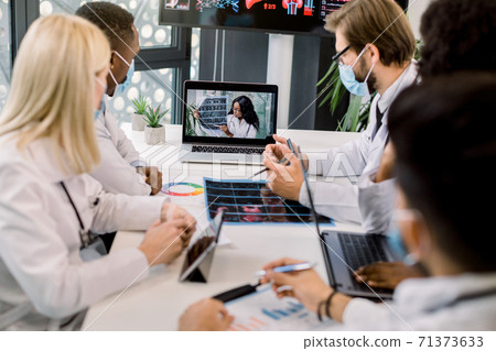 Team of multiracial experienced doctors in facial masks, having video conference with their young confident African female expert and talking about the results of patient's CT during clinical trial Team of multiracial experienced doctors in facial masks, having video conference with their young confident African female expert and talking about the results of patient's CT during clinical trial 71373633