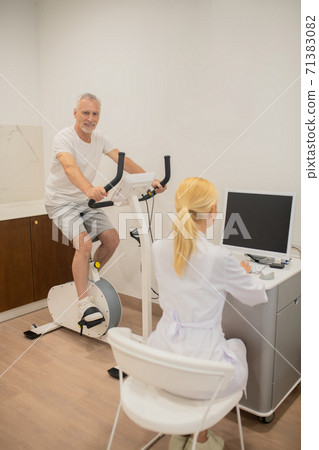 Blonde doctor in lab coat sitting at the laptop while her patient exercising on a treadmill Blonde doctor in lab coat sitting at the laptop while her patient exercising on a treadmill 71383082