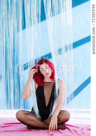 Full body vertical frame with young woman with red appearance in a black body sits on in the middle of shiny curtain against a blue wall and with a bored expression corrects hair withhand looking down 71383549