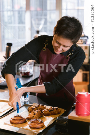 Woman chef baker barista in apron during process of glaze buns with caramel. Smiley female person with short brunette hair cut preparing pastry in an authentic coffee shop interior. 71383564