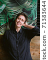 Vertical crop portrait of real woman standing indoor in front of green tropical interrior wall dreamily fixes her hair with her hand, looking straight at the camera friendly 71383644