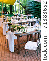 The rows of beautifully served wooden festive tables are arranged in a clear order, covered with fabric tablecloths, decorated with flower bouquets and filled with glasses and utensils.  71383652