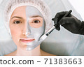 Nice woman looks directly into the camera while lying on a cosmetology table and applying a silver beauty mask to her skin with a brush. Skin cleansing and rejuvenation concept 71383663