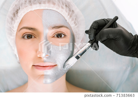 Nice woman looks directly into the camera while lying on a cosmetology table and applying a silver beauty mask to her skin with a brush. Skin cleansing and rejuvenation concept 71383663