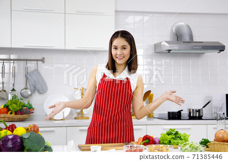 Happy Asian woman with red apron standing in the kitchen. Concept woman cooking at home. Happy Asian woman with red apron standing in the kitchen. Concept woman cooking at home. 71385446