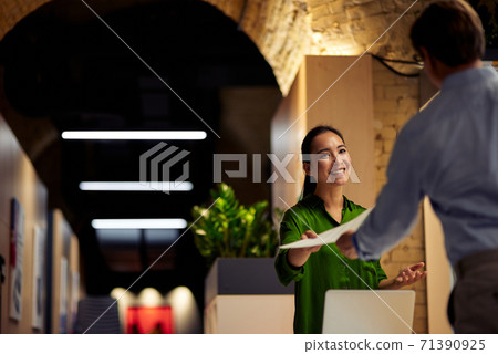 Young cheerful asian woman giving some documents to her colleague and smiling while standing together in the modern office Young cheerful asian woman giving some documents to her colleague and smiling while standing together in the modern office 71390925