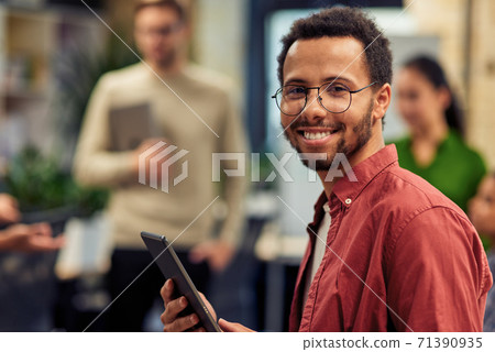 Young cheerful mixed race man holding digital tablet and smiling at camera while standing in the modern office with his colleagues on the background Young cheerful mixed race man holding digital tablet and smiling at camera while standing in the modern office with his colleagues on the background 71390935