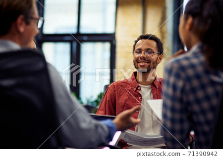 Young smiling mixed race male office worker sitting at the desk and talking with colleagues, working together in the modern office 71390942