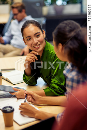 Vertical shot of a young cheerful female office worker discussing something with her colleague while sitting at the desk and working together in coworking space or office 71390955