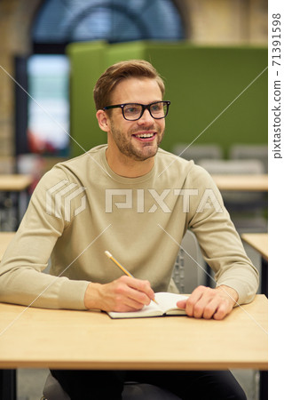Cheerful male office worker sitting at desk in coworking space, making some notes and smiling during corporate training or seminar Cheerful male office worker sitting at desk in coworking space, making some notes and smiling during corporate training or seminar 71391598