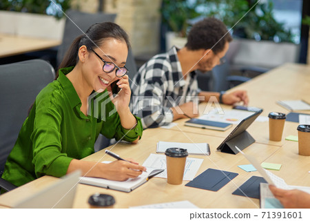 Young cheerful asian woman talking by phone and making some notes while working in the modern office Young cheerful asian woman talking by phone and making some notes while working in the modern office 71391601