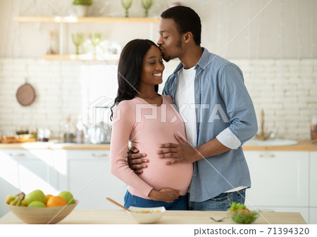 Maternity and healthy eating concept. Caring black husband kissing his pregnant wife in kitchen while cooking breakfast 71394320